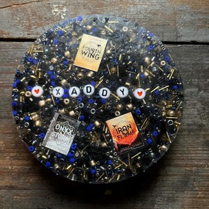 May include: A round resin coaster with black, blue, and gold beads, featuring the words "XADDY" with hearts on either side. The coaster also includes book covers for "Onyx and Storm" and "Iron and Flame" by Rebecca Yarros, and a small square sticker that says "Fourth Wing" by Rebecca Yarros.