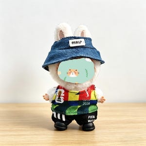May include: A plush bunny toy wearing a denim bucket hat with "MRUP" text, a colorful soccer jersey with "2026" and "FIFA World Cup" text, and black shorts. The bunny has a cat face on its face.