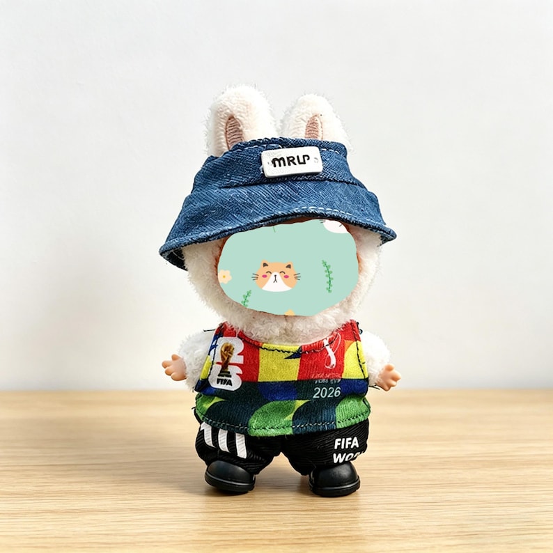 May include: A plush bunny toy wearing a denim bucket hat with "MRUP" text, a colorful soccer jersey with "2026" and "FIFA World Cup" text, and black shorts. The bunny has a cat face on its face.