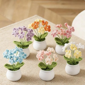 Puede incluir: Una colección de seis pequeños arreglos florales de ganchillo hechos a mano en macetas de cerámica blancas. Las flores son de varios colores, incluyendo azul, morado, naranja, rosa y amarillo, con hojas verdes. Cada arreglo es único.