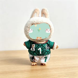 May include: A plush bunny doll wearing a green and white soccer uniform with the number 1. The doll has a fluffy white head and ears, and the face is covered with a light green patch with cat faces. The uniform has the number 1 on the front and back.