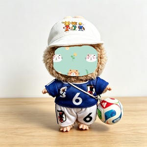 May include: A plush toy wearing a white cap with cartoon characters, a blue soccer jersey with the number 6, and white shorts. The toy is holding a soccer ball with red, blue, and white panels. The toy has a light brown fur.
