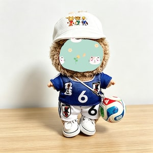 May include: A plush teddy bear wearing a white baseball cap with a cartoon design, a blue soccer jersey with the number 6, white shorts, and white sneakers. The bear is holding a miniature soccer ball.