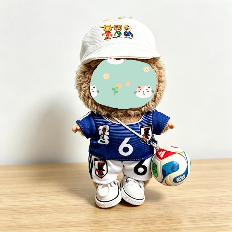 May include: A plush teddy bear wearing a white baseball cap with a cartoon design, a blue soccer jersey with the number 6, white shorts, and white sneakers. The bear is holding a miniature soccer ball.