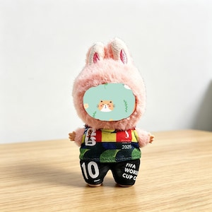 May include: A plush toy bunny wearing a soccer uniform. The bunny has a pink, fluffy head with upright ears and a face shield with a cat design. The uniform is green, black, red, and yellow with the number 10 and the text "FIFA WORLD CUP C" on the shorts.