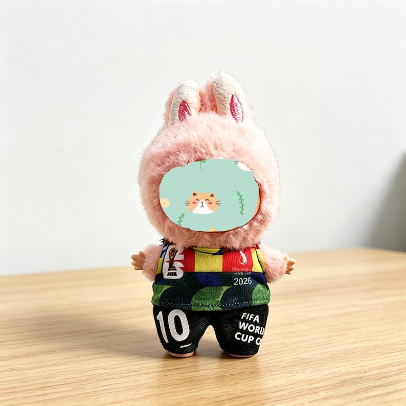 May include: A plush toy bunny wearing a soccer uniform. The bunny has a pink, fluffy head with upright ears and a face shield with a cat design. The uniform is green, black, red, and yellow with the number 10 and the text "FIFA WORLD CUP C" on the shorts.
