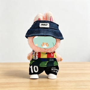 May include: A plush pink bunny doll wearing a denim hat with "MRU" text, a colorful jersey with "FIFA WORLD CUP 2026" text, black shorts with the number "10", and white shoes. The doll has a cat face.