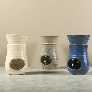 May include: Three ceramic oil warmers in cream, gray, and blue. Each warmer has a bulbous base with an oval opening and a separate dish on top. The cream warmer is on the left, the gray in the middle, and the blue on the right.