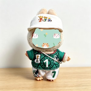 May include: A plush toy rabbit wearing a white baseball cap with a cartoon design, a green soccer jersey with the number 1, and white shorts with the number 7. The rabbit has a light brown face and ears.