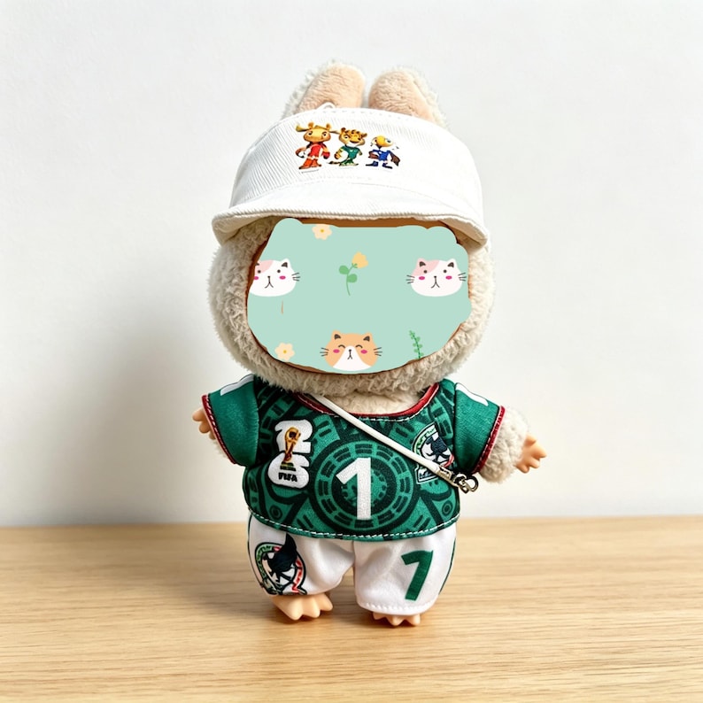 May include: A plush toy rabbit wearing a white baseball cap with a cartoon design, a green soccer jersey with the number 1, and white shorts with the number 7. The rabbit has a light brown face and ears.