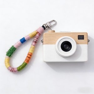 Könnte beinhalten: Eine Spielzeugkamera aus Holz mit weißem Gehäuse und hellbraunem Oberteil. Die Kamera hat ein schwarzes Objektiv und einen kleinen schwarzen Sucher. Eine farbenfrohe Handschlaufe mit silbernem Verschluss ist an der Kamera befestigt. Der Riemen besteht aus Garn in verschiedenen Farben.
