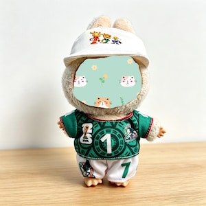 May include: A plush toy bunny wearing a white baseball cap with cartoon figures and a green soccer jersey with the number 1 and the Mexican flag colors. The bunny is also wearing white shorts with the number 7.