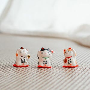 Może przedstawiać: Trzy ceramiczne figurki Maneki Neko. Każdy kot ma białe ciało z czarnymi, czerwonymi i pomarańczowymi akcentami. Znajdują się na małych czerwonych podstawach i zawierają japońskie znaki. Jeden kot trzyma złotą monetę, inny ma podniesioną łapę, a trzeci ma podniesioną łapę.
