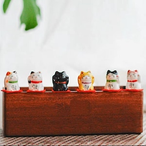 May include: A collection of six ceramic Maneki Neko figurines in various colors, including white, black, and orange, each with unique details. They sit on a wooden base with red mats, embodying the traditional Japanese lucky cat theme.