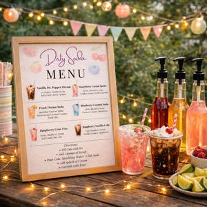 May include: A framed Dirty Soda menu sign with illustrations of fruit, listing drink recipes like Vanilla Dr. Pepper Dream and Peach Dream Soda. Next to the sign are three syrup bottles with pumps and two clear plastic cups filled with iced drinks.