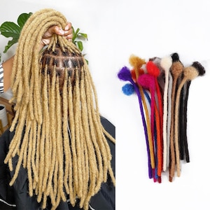 Puede incluir: Cabeza de una persona con rastas rubias, sostenida por una mano. A la derecha, una colección de extensiones de rastas coloridas en varios colores, incluyendo azul, morado, rojo y marrón. Las extensiones están hechas de un material suave y difuso.