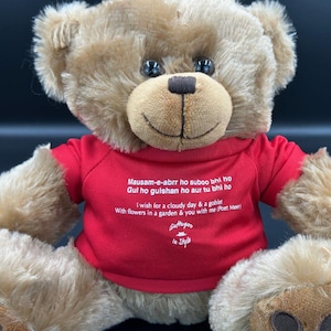 May include: A brown teddy bear wearing a red shirt with white text that reads "Mausam-e-abrr ho suboo bhi ho Gul ho gulshan ho aur tu bhi ho I wish for a cloudy day & a goblet With flowers in a garden & you with me (Poet Meer) Guttugoo In Style"