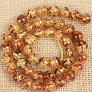 May include: A strand of amber-colored beads with a mix of light and dark brown inclusions. The round beads are strung together on a yellow string, creating a necklace or bracelet component. The beads have a polished, glossy finish.