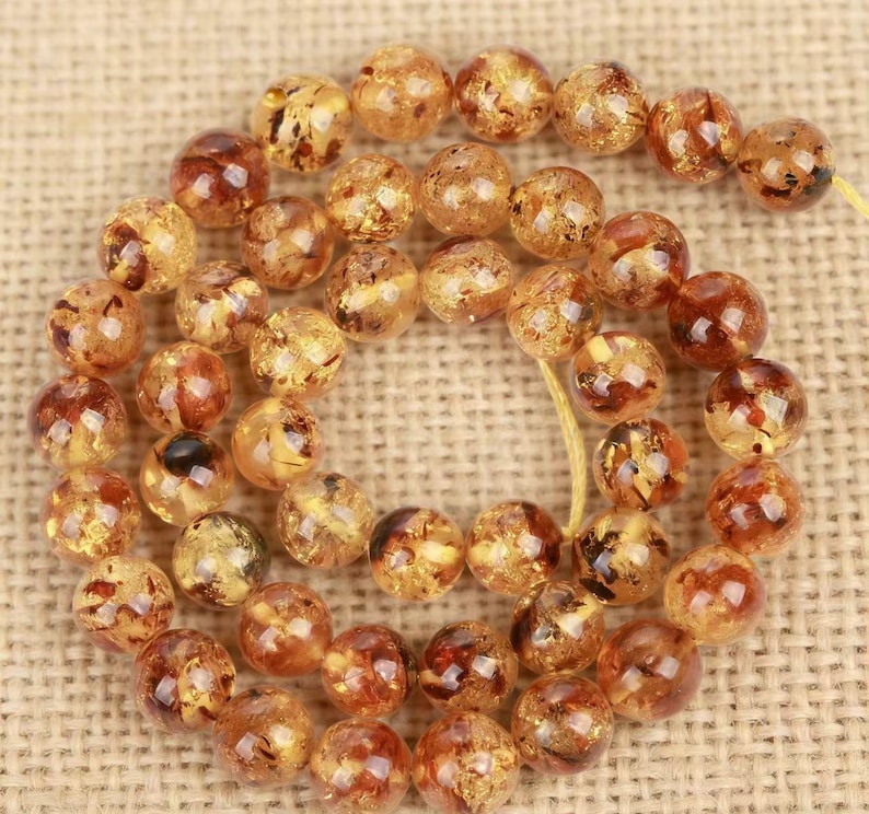 May include: A strand of amber-colored beads with a mix of light and dark brown inclusions. The round beads are strung together on a yellow string, creating a necklace or bracelet component. The beads have a polished, glossy finish.