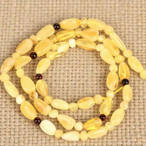 May include: A necklace composed of light yellow amber beads in various shapes, including oval and teardrop. The necklace features small, dark brown accent beads and is displayed on a textured, tan background. The amber beads have a translucent quality.