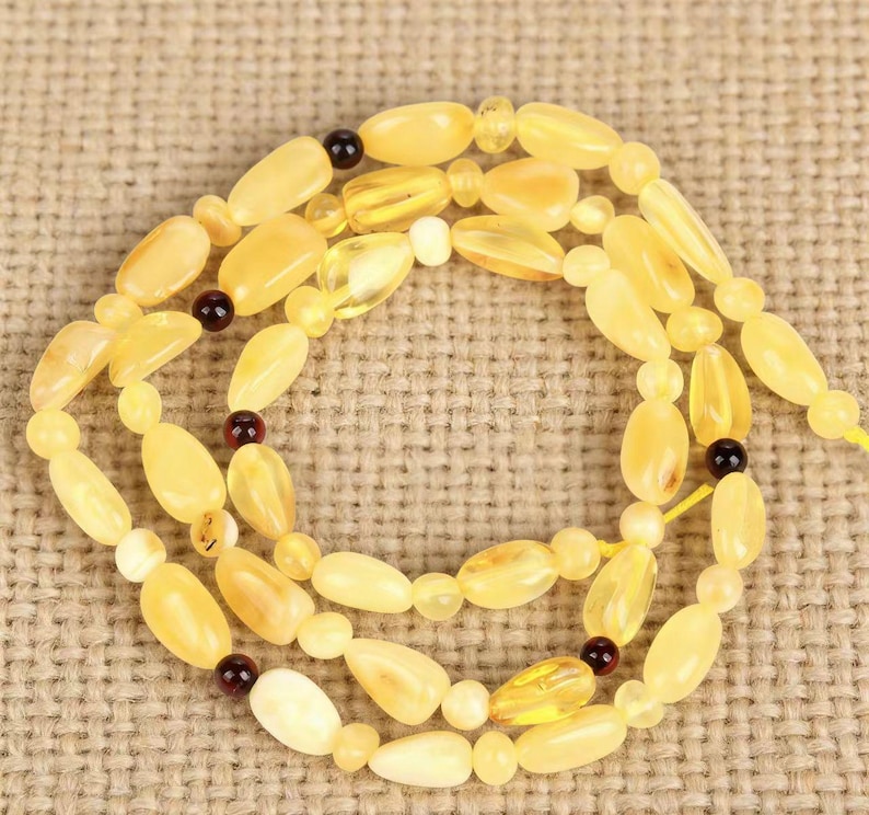 May include: A necklace composed of light yellow amber beads in various shapes, including oval and teardrop. The necklace features small, dark brown accent beads and is displayed on a textured, tan background. The amber beads have a translucent quality.