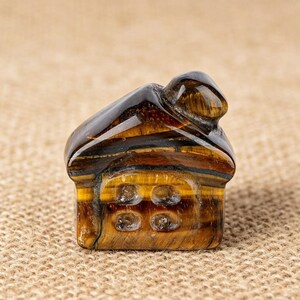 May include: A small, intricately carved house figurine made from tiger's eye gemstone. The house features a dark brown roof and a golden-brown body with four window details. The gemstone has natural bands of color.