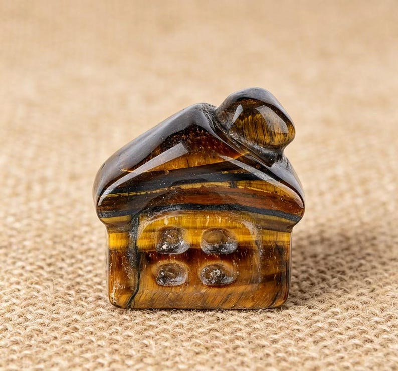 May include: A small, intricately carved house figurine made from tiger's eye gemstone. The house features a dark brown roof and a golden-brown body with four window details. The gemstone has natural bands of color.