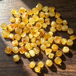 May include: A collection of numerous small, polished amber beads in various shades of yellow, orange, and honey brown. Each bead features a smooth, rounded, or irregular shape with a central hole, displaying natural variations in colour and translucency.