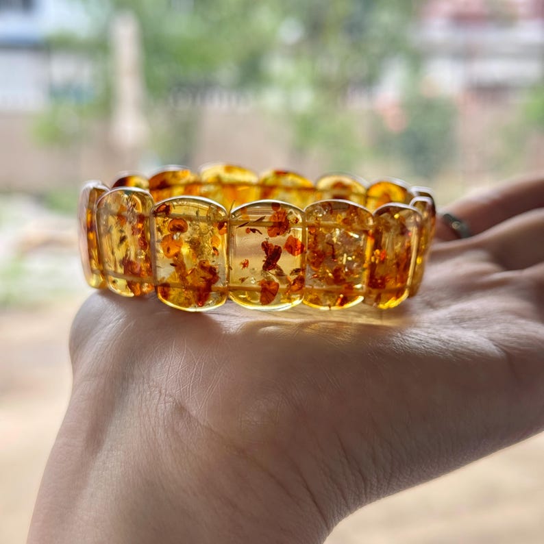 May include: A translucent amber bracelet with rectangular segments. The bracelet has a warm, golden-yellow hue with reddish-brown inclusions. The bracelet is shown on a person's hand, with a blurred background of greenery.