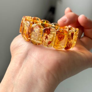 May include: A chunky amber bracelet with rectangular, translucent beads. The beads have a warm, golden-yellow hue with reddish-brown inclusions, creating a unique, natural pattern. The bracelet is held in a hand, showcasing its size and design.