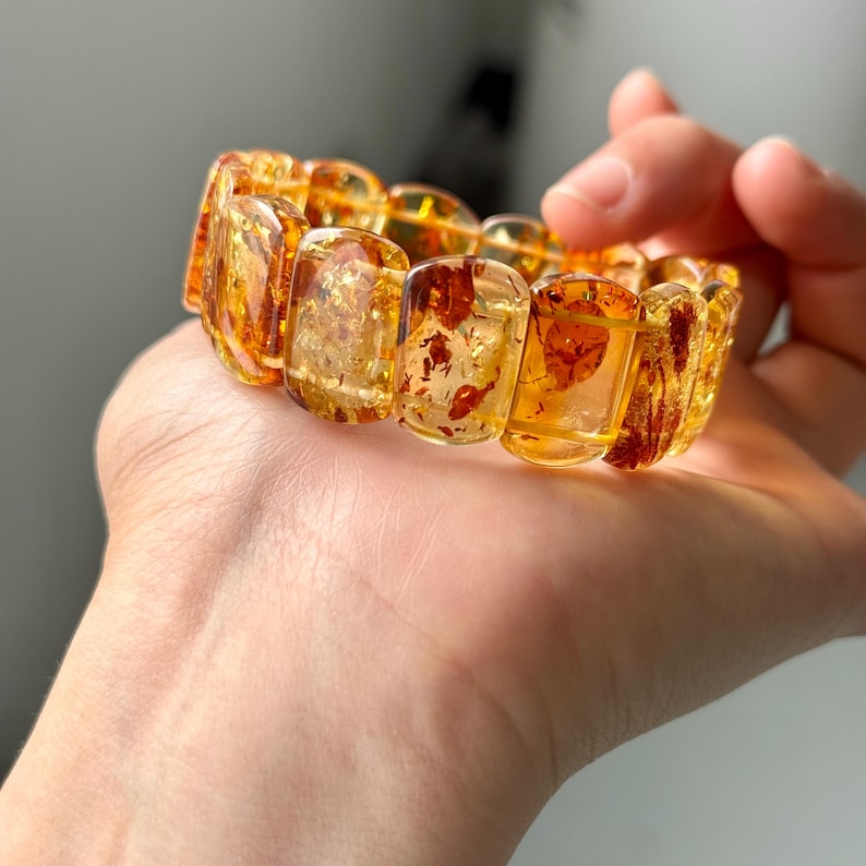 May include: A chunky amber bracelet with rectangular, translucent beads. The beads have a warm, golden-yellow hue with reddish-brown inclusions, creating a unique, natural pattern. The bracelet is held in a hand, showcasing its size and design.