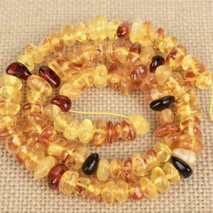 May include: A necklace of polished amber beads in various shades of yellow, brown, and black. The beads are strung together on a yellow string, creating a spiral pattern. The amber beads have an irregular shape and a glossy finish.