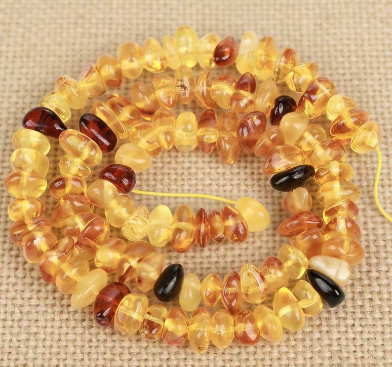 May include: A necklace of polished amber beads in various shades of yellow, brown, and black. The beads are strung together on a yellow string, creating a spiral pattern. The amber beads have an irregular shape and a glossy finish.