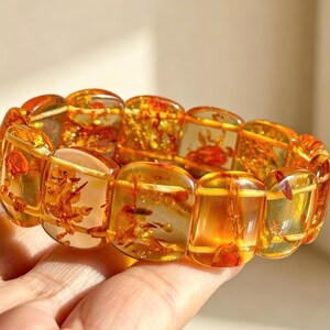 May include: A bracelet featuring large, rectangular amber beads. The translucent beads display warm orange and yellow hues, with visible inclusions. The bracelet is held in a hand, showcasing the polished, natural beauty of the amber.