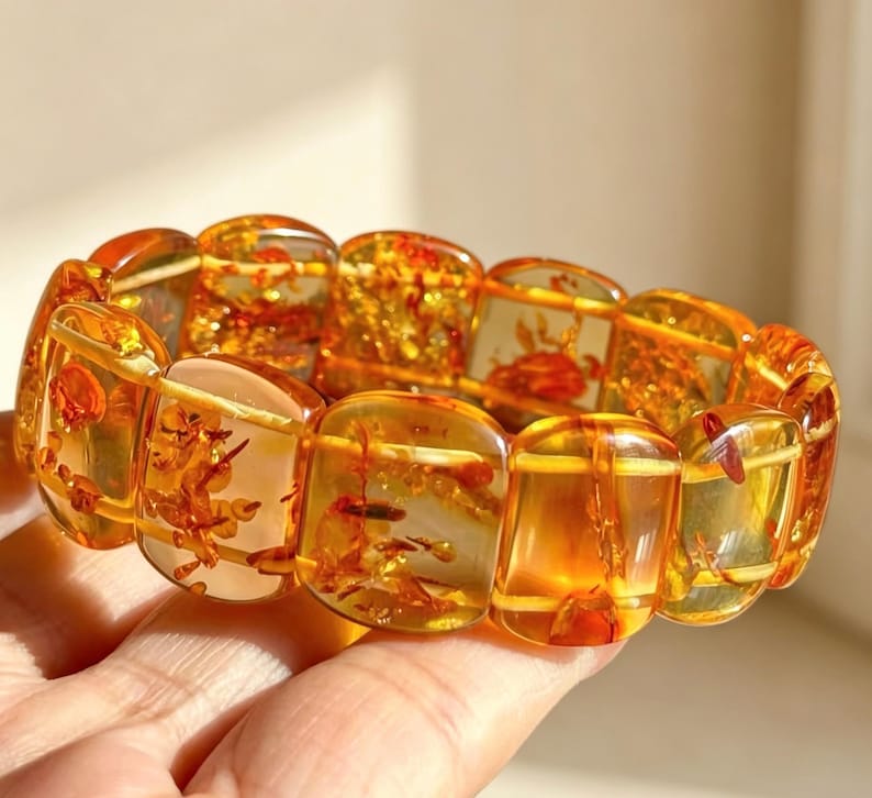 May include: A bracelet featuring large, rectangular amber beads. The translucent beads display warm orange and yellow hues, with visible inclusions. The bracelet is held in a hand, showcasing the polished, natural beauty of the amber.