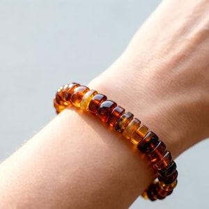 May include: A stretch bracelet composed of numerous polished, rounded amber-colored beads. The beads exhibit varying shades of translucent orange, golden yellow, and deep brown, creating a warm, multi-tonal appearance around the wrist.