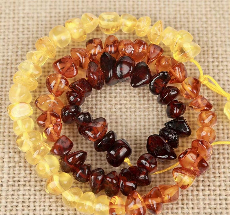 May include: Strands of amber gemstone beads in varying shades of yellow, orange, and brown. The beads are irregularly shaped and strung together, creating a circular pattern. The background is a neutral tan fabric.
