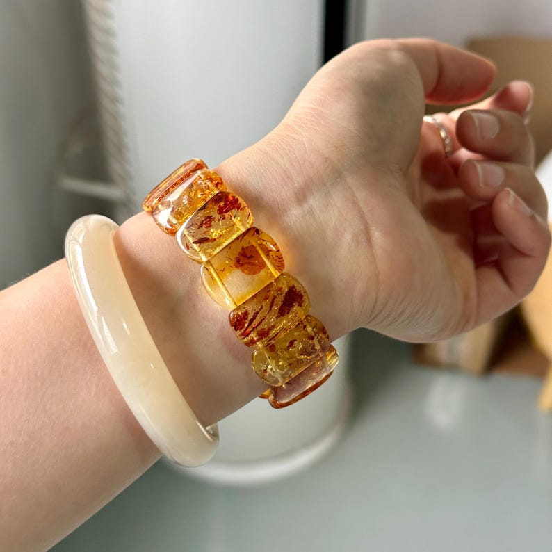 May include: A wrist adorned with two bracelets. One is a light cream-colored bangle. The other is a bracelet made of amber-colored, square-shaped beads. The amber beads have a warm, translucent quality with reddish-brown inclusions.