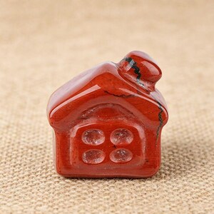 May include: A small, red jasper house figurine. The miniature house has a pitched roof, a chimney, and a square base with four circular window details. The polished stone has natural variations in color and texture, adding to its unique charm.