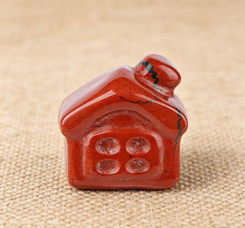 May include: A small, red jasper house figurine. The miniature house has a pitched roof, a chimney, and a square base with four circular window details. The polished stone has natural variations in color and texture, adding to its unique charm.