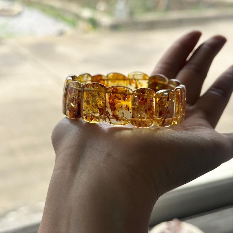 May include: A translucent amber bracelet with rectangular segments. The bracelet has a warm, golden hue with reddish-brown inclusions. The bracelet is held in a hand, showcasing its color and design.