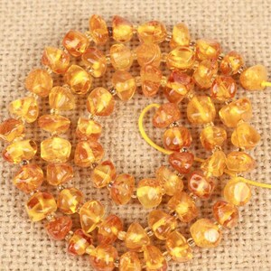 May include: A strand of polished, irregular amber beads in various shades of golden yellow and orange. The translucent, rounded stones are strung together on a thin yellow cord, creating a circular arrangement of natural resin pieces.