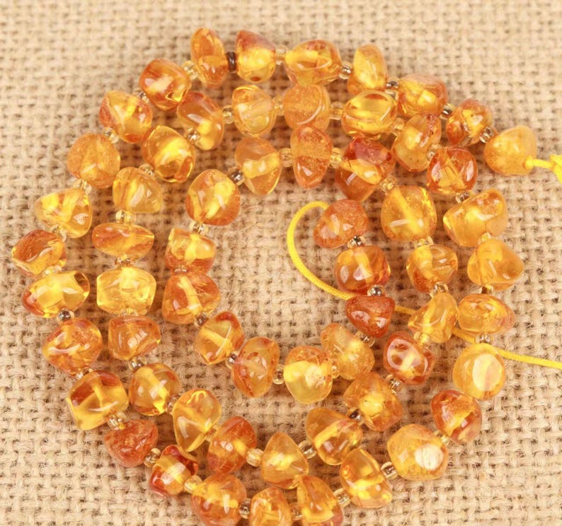 May include: A strand of polished, irregular amber beads in various shades of golden yellow and orange. The translucent, rounded stones are strung together on a thin yellow cord, creating a circular arrangement of natural resin pieces.