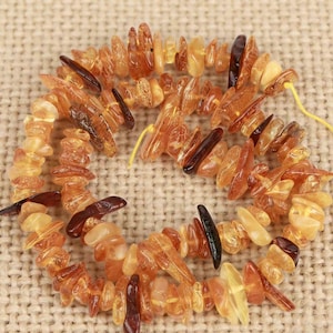 May include: A strand of irregular, tumbled amber beads in various shades of translucent honey, golden yellow, and deep reddish-brown. The polished, organic-shaped stones are strung together on a thin yellow cord, creating a textured necklace.