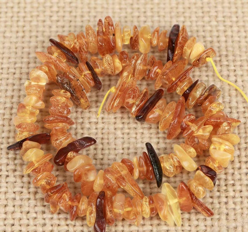 May include: A strand of irregular, tumbled amber beads in various shades of translucent honey, golden yellow, and deep reddish-brown. The polished, organic-shaped stones are strung together on a thin yellow cord, creating a textured necklace.