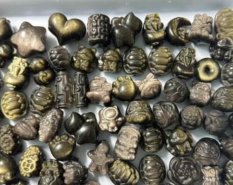 May include: A collection of small, carved obsidian beads with a metallic gold sheen, featuring various shapes including stars, hearts, turtles, and animal figures, arranged together on a white tray for jewelry making and crafting projects.