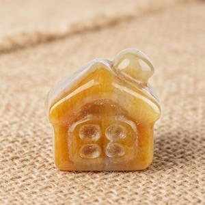 May include: A small, house-shaped figurine carved from a translucent, golden-yellow stone. The miniature house features a rounded roof and a front facade with four circular window details. The stone has natural variations in color and texture.