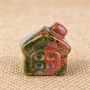 May include: A small, carved stone house figurine. The house is a mix of red and green colors, with a rounded roof and two oval-shaped windows. The stone has a polished, smooth surface.
