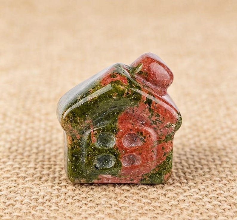 May include: A small, carved stone house figurine. The house is a mix of red and green colors, with a rounded roof and two oval-shaped windows. The stone has a polished, smooth surface.