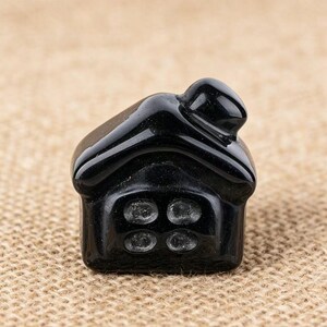 May include: A small, glossy black house figurine with four circular window details. The roof has a curved design, and the overall shape is compact and detailed. The figurine is set against a neutral, textured background.